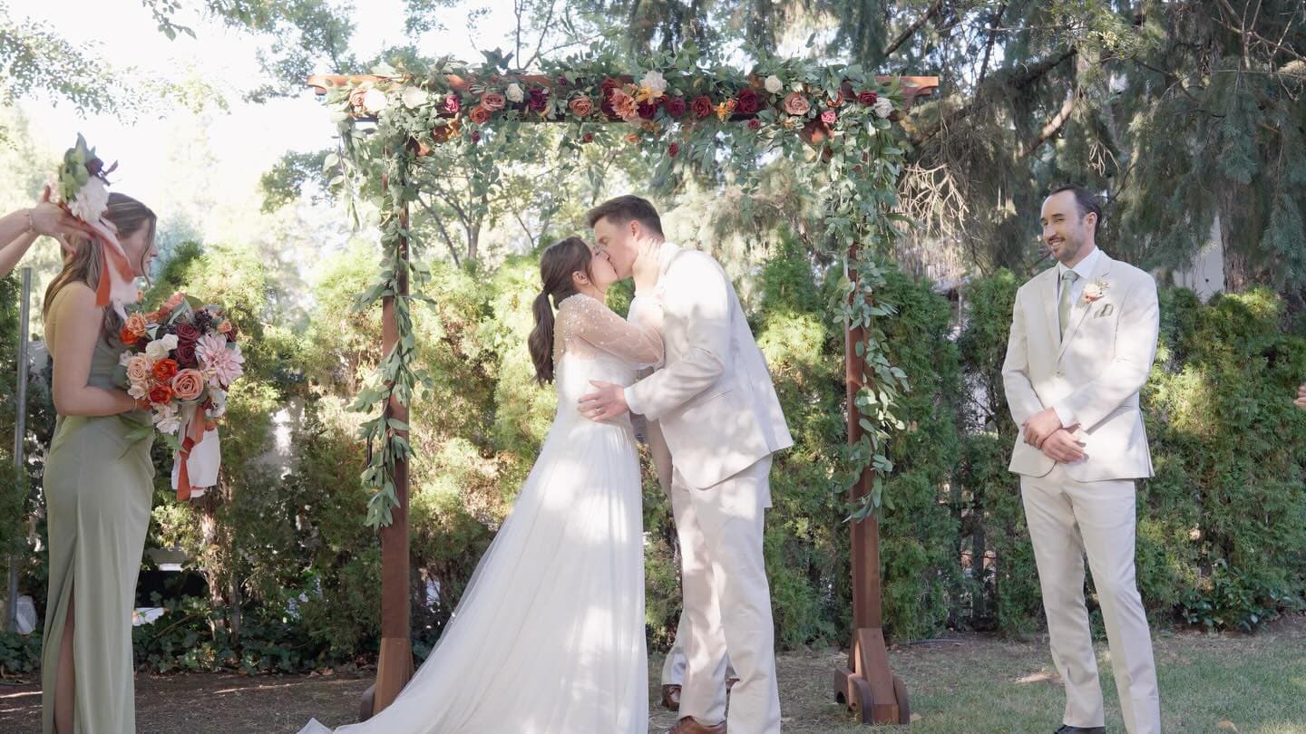 A couple exchanges a kiss under a floral wedding arch, surrounded by guests.