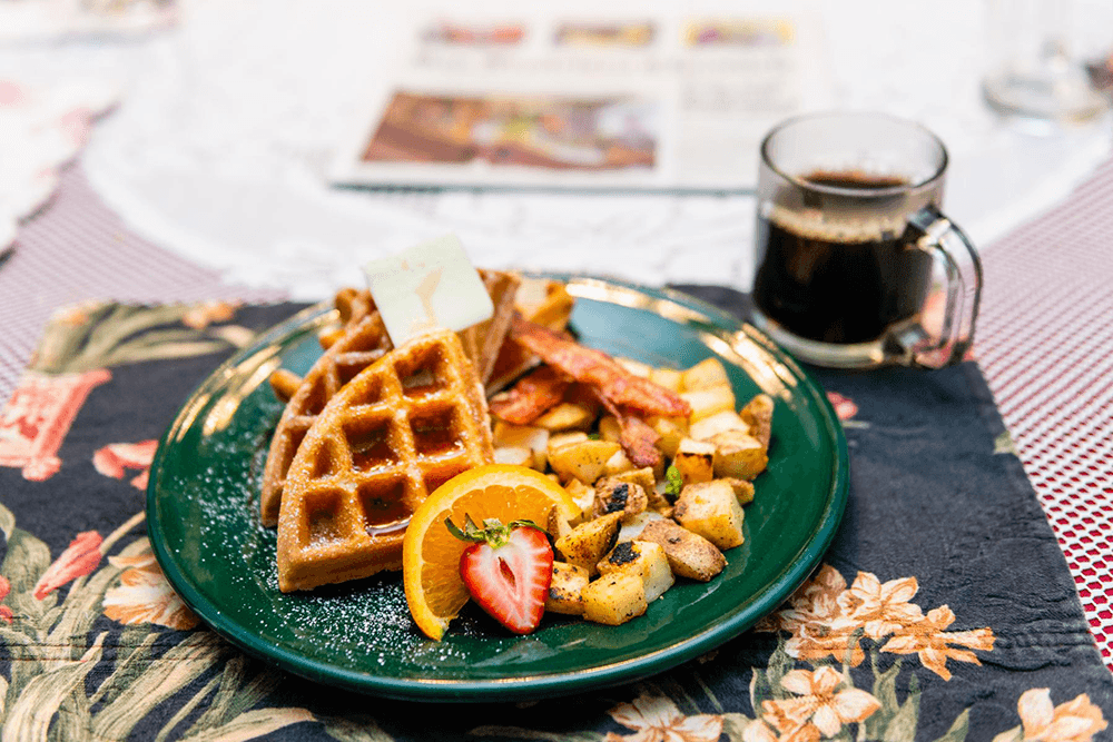 A green plate holding waffles, bacon, and golden-brown potatoes, garnished with a slice of orange and a strawberry, alongside a cup of coffee.
