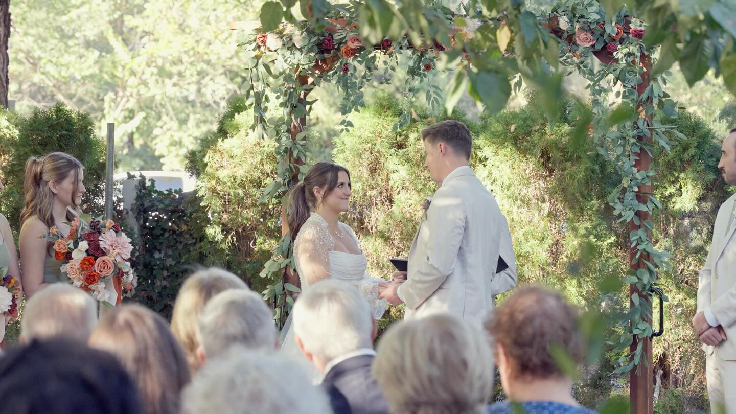 A bride and groom exchange vows under a floral arch at an outdoor wedding ceremony.