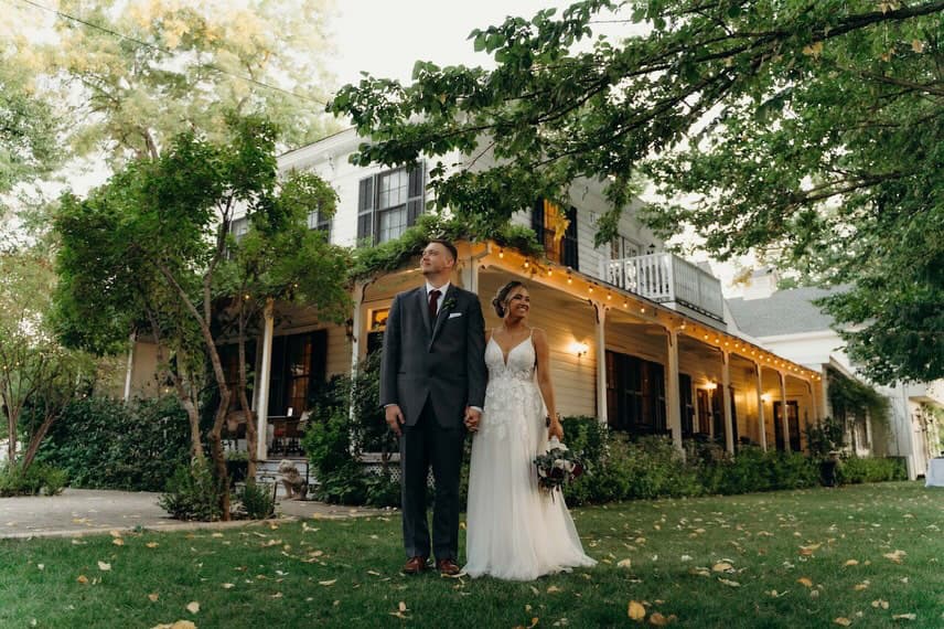 A newlywed couple stands hand in hand in front of a charming house adorned with outdoor lights.