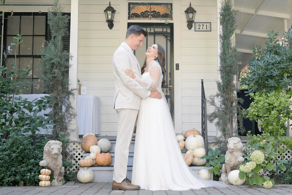 A couple in formal attire embraces joyfully on a porch adorned with pumpkins.