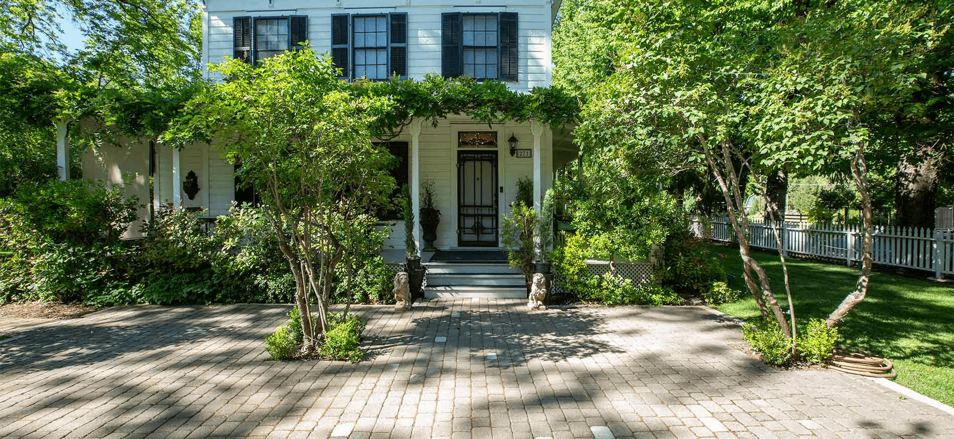A charming white house surrounded by lush greenery and a brick pathway.